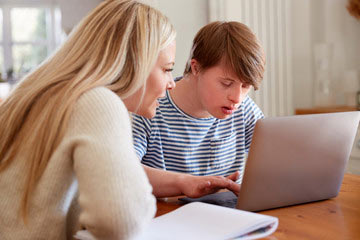 A woman tutoring a young man as he types on his laptop computer at the kitchen table