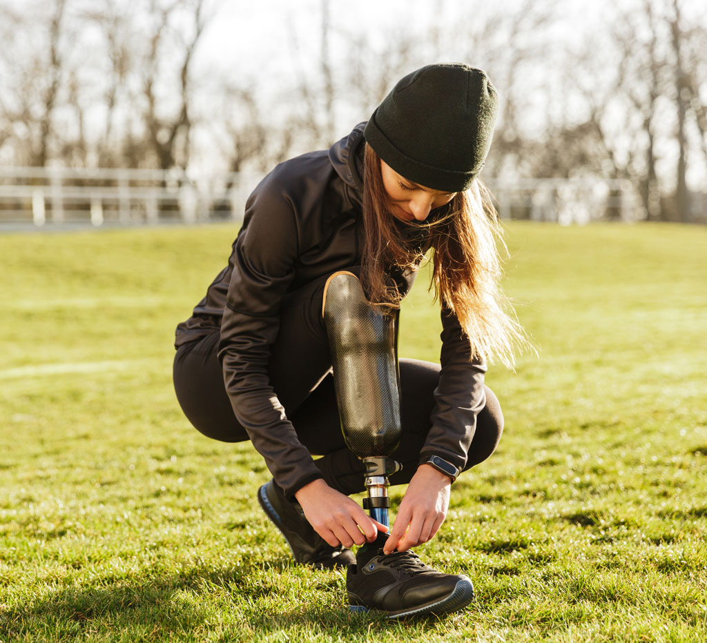 a woman in black athletic wear and black beanie tying her shoe at the end of her prosthetic leg and foot