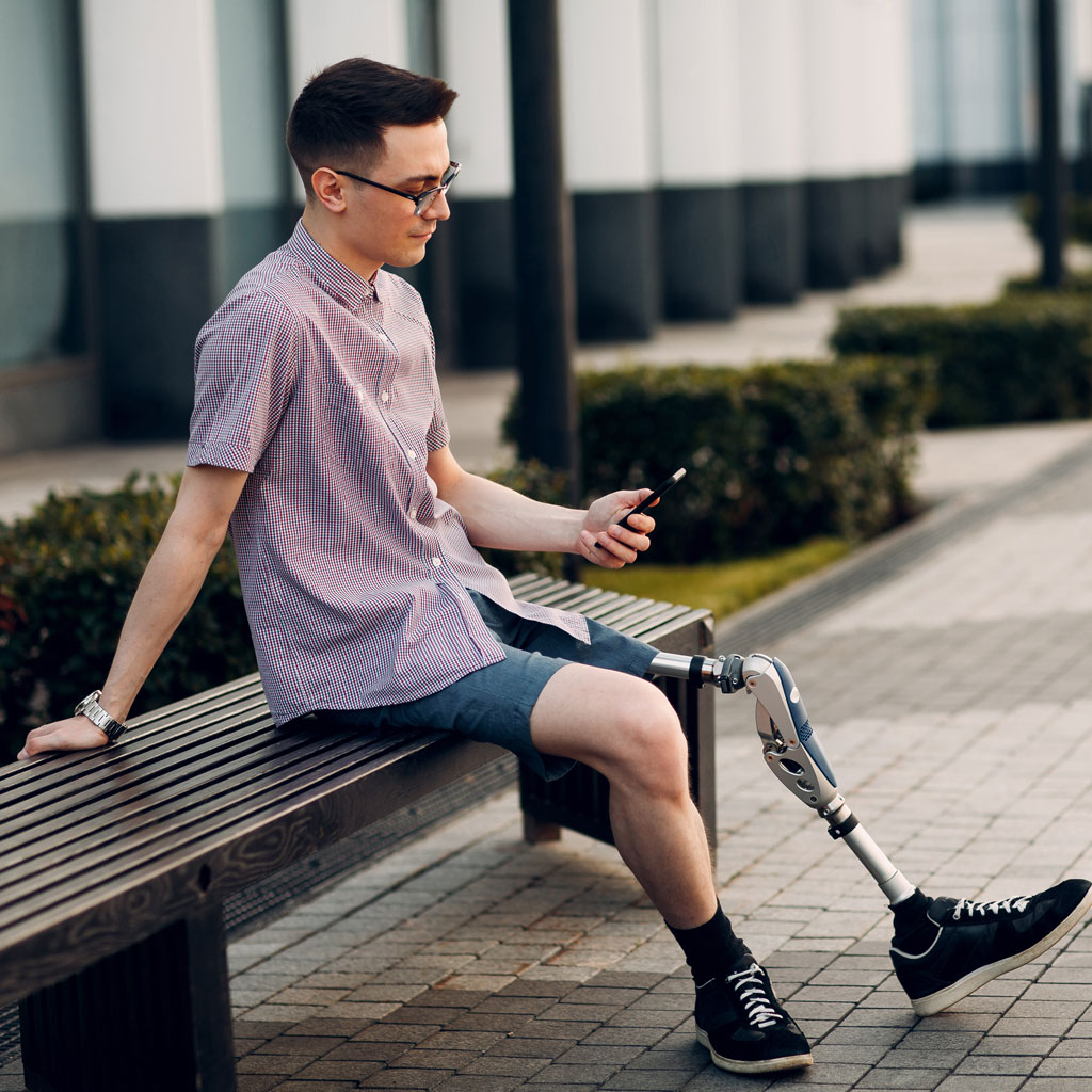 man with prosthetic leg sitting on a park bench outside whilst looking at his phone